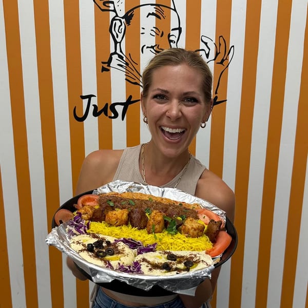 Smiling woman holding a large platter of Middle Eastern food with rice, kebabs, hummus, and vegetables.