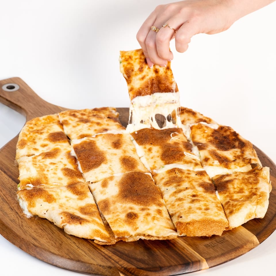 A hand pulls a cheesy slice of Middle Eastern flatbread from a wooden board, stretching the melted cheese.