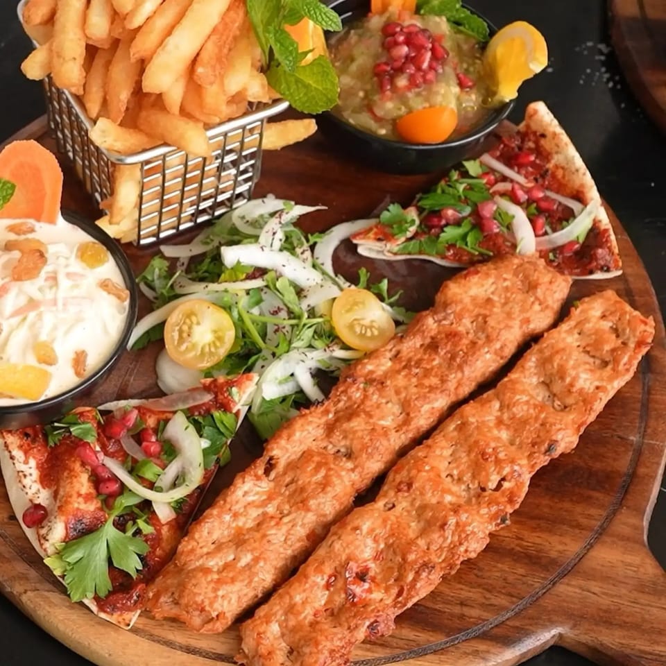 Middle-Eastern platter with two kebabs, fries, dips, salad, and flatbread on a wooden board.