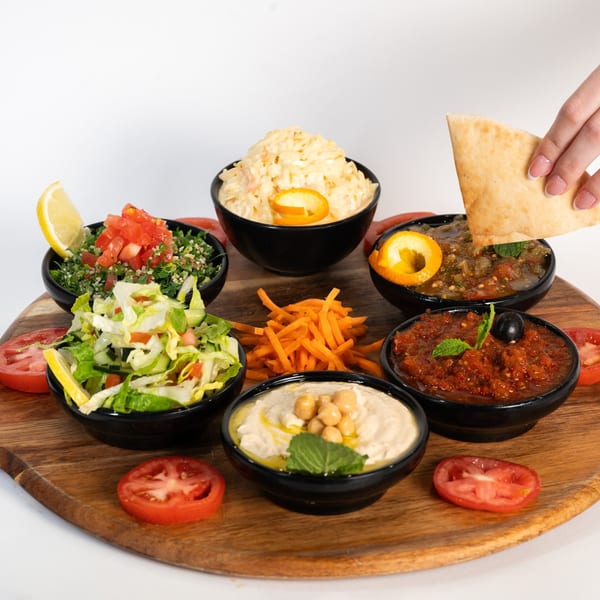A wooden platter with six bowls of Middle Eastern dips and salads, with a hand dipping pita bread.