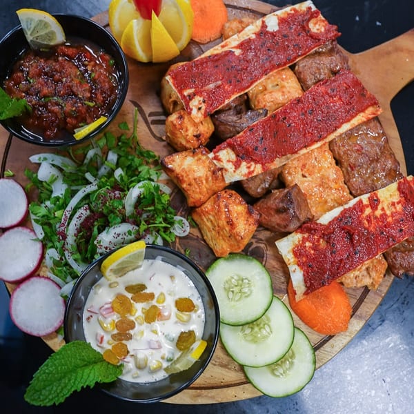 A wooden board with Middle Eastern kebabs, flatbread, salads, and dips, garnished with lemon and mint.