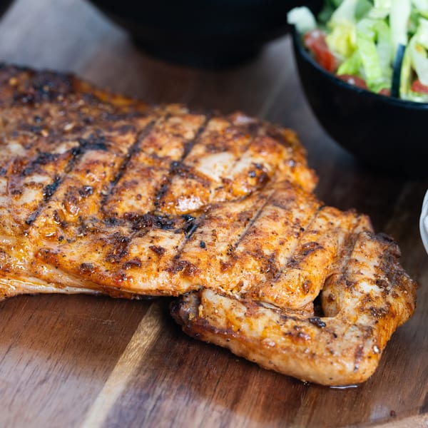 Grilled chicken with char marks on a wooden board, with a side salad in a black bowl.