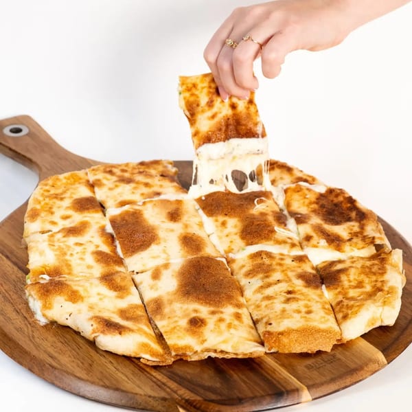 A hand pulls a cheesy slice of Middle Eastern flatbread from a wooden board, stretching the cheese.