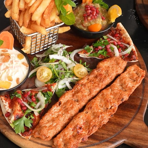 Middle-eastern platter with kebab, fries, dips, salad, and flatbread on a wooden board.