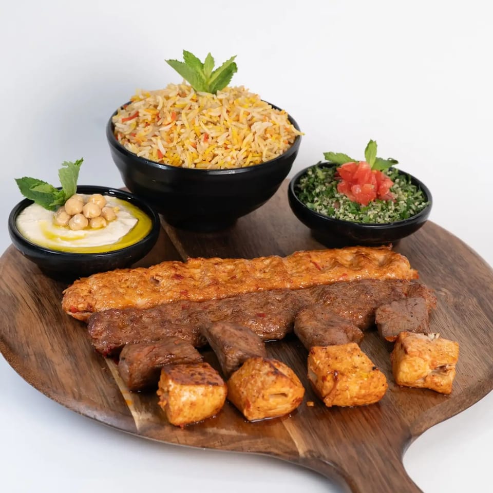 Middle-Eastern platter with various grilled meats, rice, hummus, and tabbouleh on a wooden board.