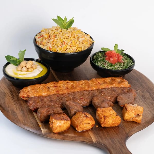 Middle-eastern platter with rice, hummus, tabbouleh, and various grilled meats on a wooden board.