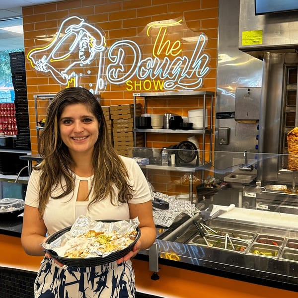 Smiling woman holding a Middle-Eastern dish in a basket, with "The Dough Show" neon sign behind her.