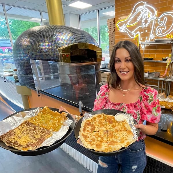A woman holds two trays of Middle Eastern pastries, one sweet and one savory, in front of a large oven.