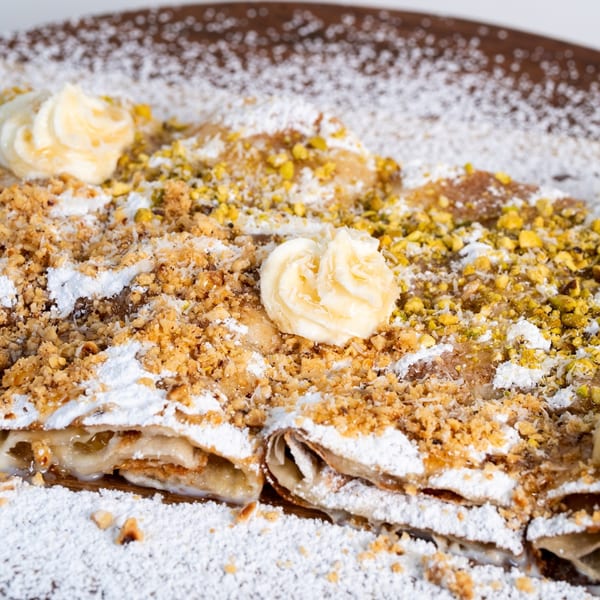 Middle-eastern dessert with powdered sugar, crushed nuts, pistachios, and cream on a brown plate.
